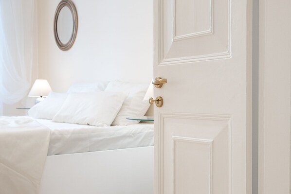 A white room with a bed, a bedside table with a lamp, and a door with a brass handle. - Corfu Charm | Chic, Central, Convenient! | Fast WiFi (Kerkira)