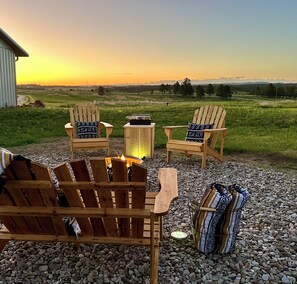 A cozy outdoor seating area with wooden Adirondack chairs and a fire pit, set against a scenic sunset view. - Serene country setting with views of the Black Hills. (Custer)