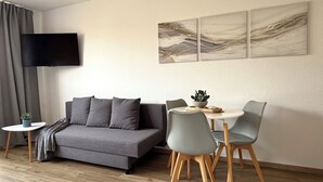 A modern living room with a grey sofa, a dining table with chairs, a flat-screen TV mounted on the wall, and a marble artwork above the table. - Luv & Lee Duhnen - Holiday apartment no. 5 near the beach (Cuxhaven)