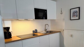 A compact kitchen with white cabinets, a black microwave, a stainless steel sink, and a white radiator. - Luv & Lee Duhnen - Holiday apartment no. 5 near the beach (Cuxhaven)