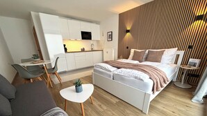 A modern bedroom with a bed, a small table with a plant, a sofa, and a kitchenette in the background. - Luv & Lee Duhnen - Holiday apartment no. 5 near the beach (Cuxhaven)