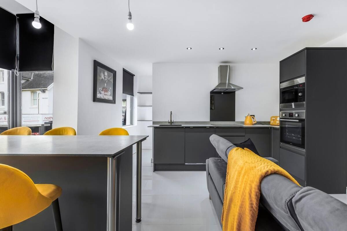 Een moderne keuken met een centraal eiland, donkere kasten en een gele plaid op een grijze bank.