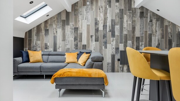 A modern loft-style living space with a sloped ceiling, built-in wooden wall paneling, a grey sectional sofa with yellow cushions, a matching ottoman, and a yellow dining chair. - Winter Garden Apartments by Sasco (Blackpool)