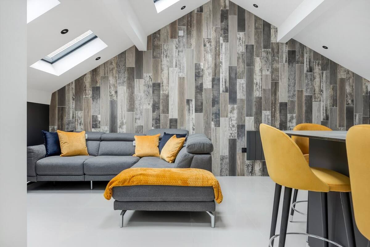 Een moderne loft-achtige woonruimte met een schuine plafond, houten wandpanelen, een grijze modulaire bank met gele kussens, een bijpassend poufje en een gele eetkamerstoel.
