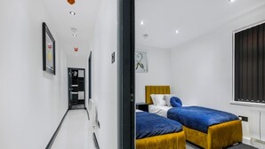 A modern hotel room with a hallway leading to a bathroom, two beds with blue and yellow bedding, and a window with blinds. - Winter Garden Apartments by Sasco (Blackpool)