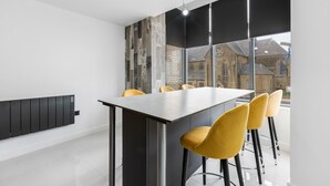 A modern kitchen with a long counter, yellow chairs, and a view of historic buildings through large windows. - Winter Garden Apartments by Sasco (Blackpool)