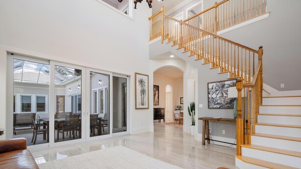 A spacious living area with a high ceiling, a wooden staircase, and a view of the backyard through large sliding glass doors. - Luxury Home in Naples with Private Pool & Walk to the Beach by Esthetic Stays (Naples)