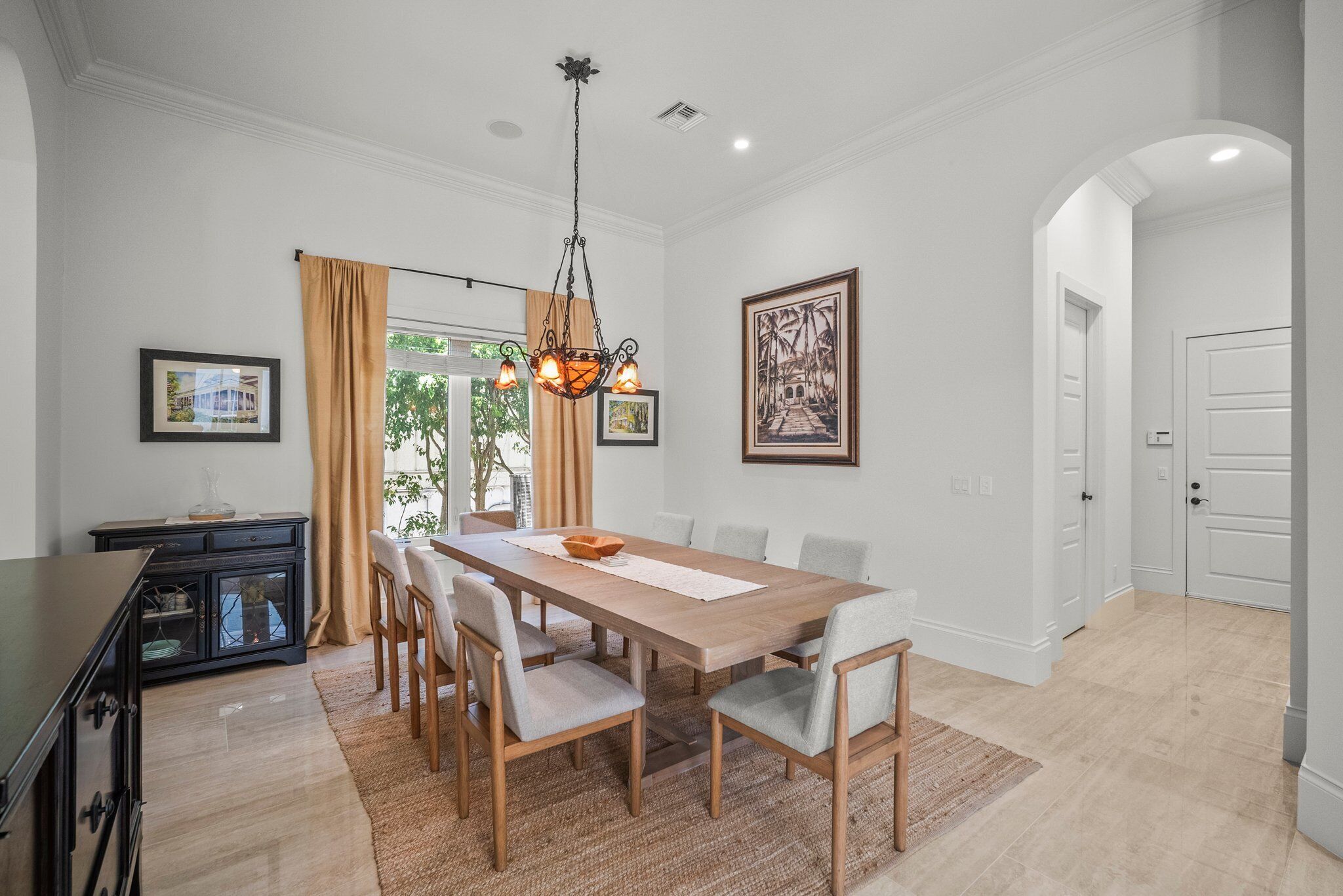 A dining area with a wooden table, six chairs, a chandelier, framed artwork, and a black cabinet.