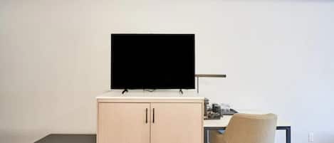 A room with a desk, a chair, a television on top of a cabinet, and a small bench.