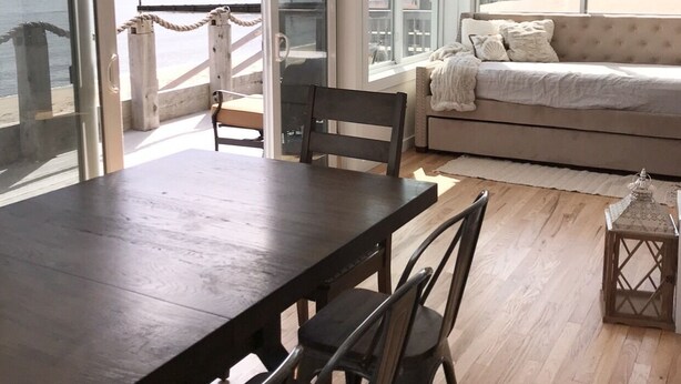 A spacious room with a large dining table and chairs, a sofa, and large windows offering a view of the sea and buildings. - Oceanfront Oasis Spacious 3-Bedroom, 2-Bath Retreat for 12  Linens Included (Old Saybrook)