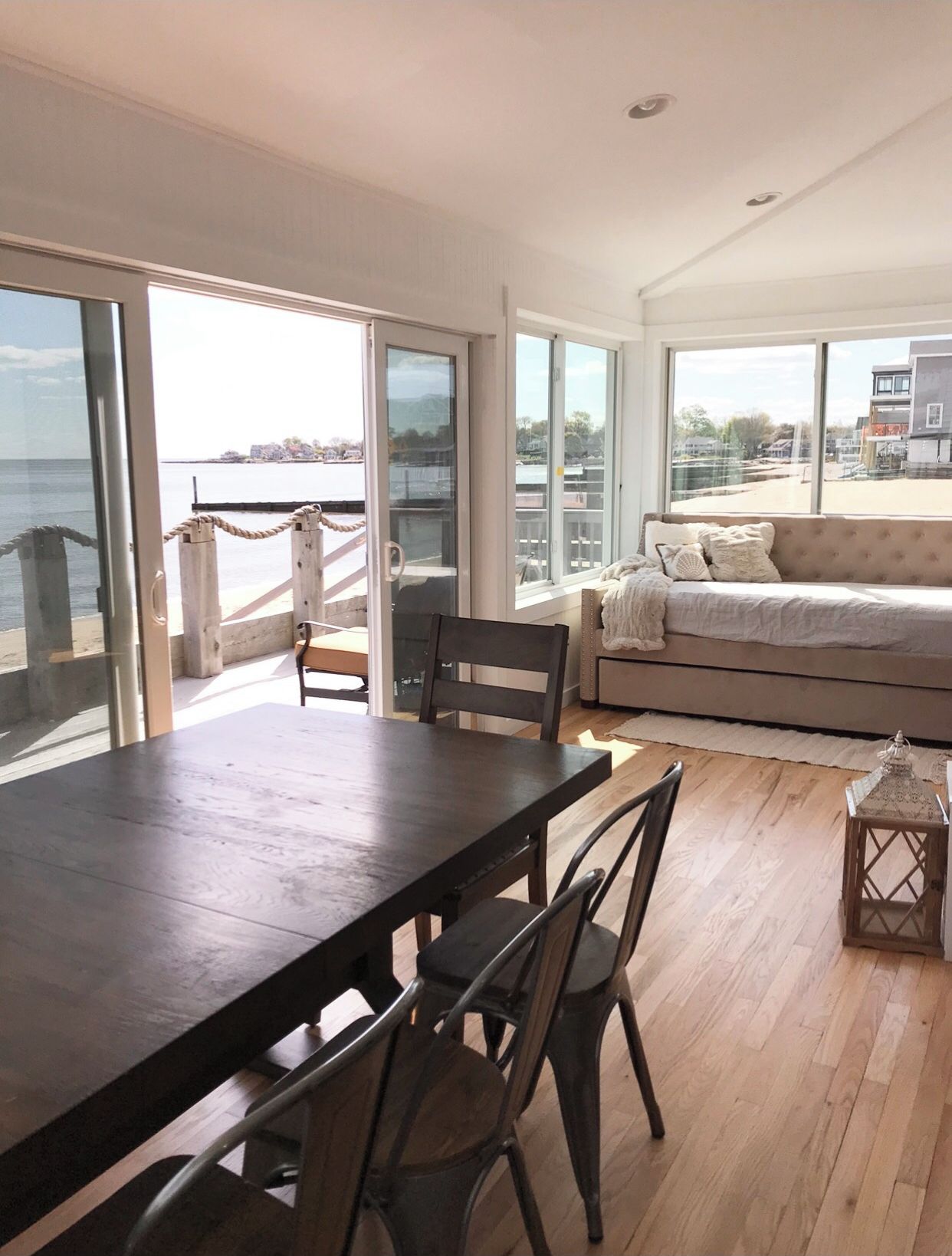 A spacious room with a large dining table and chairs, a sofa, and large windows offering a view of the sea and buildings.