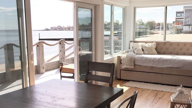 Amplia habitación con una gran mesa de comedor y sillas, un sofá, y ventanas amplias que ofrecen vista al mar y edificios.