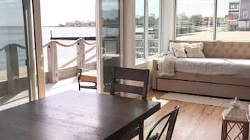 A spacious room with a large dining table and chairs, a sofa, and large windows offering a view of the sea and buildings.