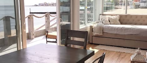 A spacious room with a large dining table and chairs, a sofa, and large windows offering a view of the sea and buildings.
