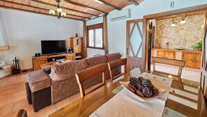 Ein gemütliches Wohnzimmer mit Holzbalkendecke, einem Fernseher, einer Couch und einem Esstisch mit Kiefernzapfen.