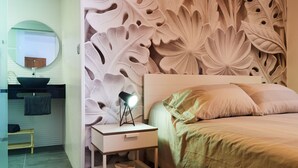 Ein Schlafzimmer mit einem Bett, einem Nachttisch mit Lampe, einem Spiegel und einer Wand mit Blättermotiv.