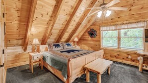 Quarto de cabana de madeira com cama, mesas de cabeceira, um banco, ventilador de teto e janela com vista para o verde.