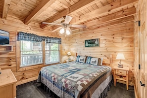 A wooden cabin bedroom with a bed, two bedside tables, a ceiling fan, and a window with blinds.