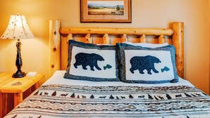 Ein Bett mit zwei Kopfkissen mit Bärenmotiv, ein Holzkopfstück, ein Nachttisch mit Lampe und ein gerahmtes Bild an der Wand.