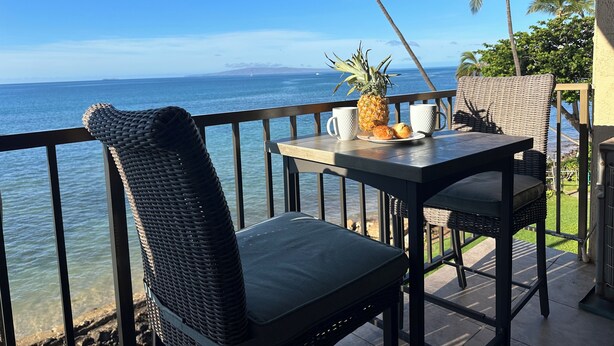 Varanda com cadeiras de vime, uma mesa com uma abacaxi e duas canecas, e vista para o oceano.