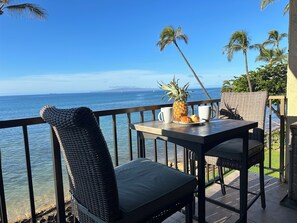 Een balkon met rieten stoelen, een tafel met een ananas en twee mokken, en uitzicht op de oceaan.