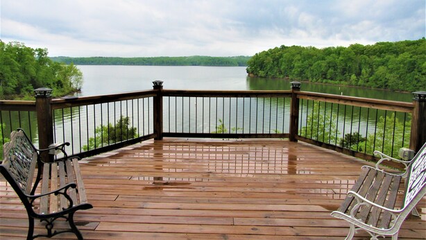 Una terraza de madera con barandilla que da a un lago, dos bancos y vistas a una orilla boscosa.