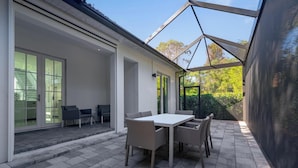 A covered patio area with a table and chairs, a staircase leading to an upper level, and a view of greenery outside. - Beautiful Naples Home with Private Pool Near Beach & Dining by Esthetic Stays (Naples)