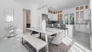 A modern kitchen with white cabinets, a central island, and a dining area with a marble table and bench. - Beautiful Naples Home with Private Pool Near Beach & Dining by Esthetic Stays (Naples)