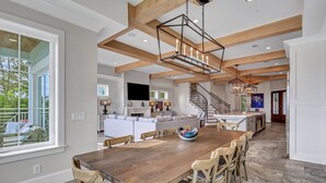 Een ruime eethoek met een lange houten tafel en stoelen, een kroonluchter, en zicht op de keuken en woonkamer op de achtergrond.