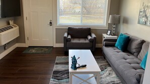 Ett vardagsrum med en grå soffa, ett vitt soffbord, en grå fåtölj, en TV som är monterad på väggen och ett fönster med persienner.