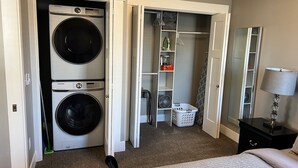 Ett sovrum med en säng, en tvättmaskin, en garderob med hyllor och ett nattduksbord med en lampa.