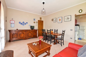 A living room with a wooden dining table, chairs, a red sofa, a coffee table with decorative items, and a kitchen area in the background.