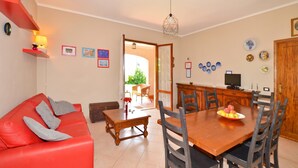 Un salon avec un canapé rouge, une table à manger en bois et des chaises, un coin cuisine avec un téléviseur, et une porte donnant sur l’extérieur.