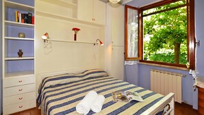 Une chambre à coucher bien rangée, avec un lit, des lampes de chevet, une étagère avec des livres et un vase, et une fenêtre donnant sur un paysage verdoyant.