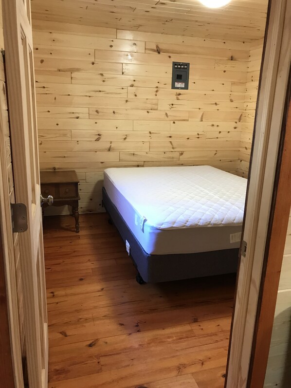 Una habitación pequeña y acogedora con paredes de madera y una cama individual. Hay una mesita de noche de madera con una lámpara y un enchufe sobre la cama.