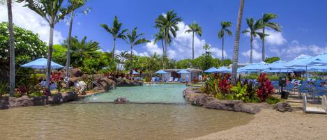 A resort pool area with clear water, surrounded by palm trees and lounge chairs under blue umbrellas.