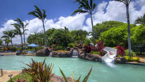 A tropical resort pool with a water slide, surrounded by lush greenery and palm trees. - Poipu Condo!  Newly updated - Sleeps 5 - 5 minute walk to the beach! (Koloa)