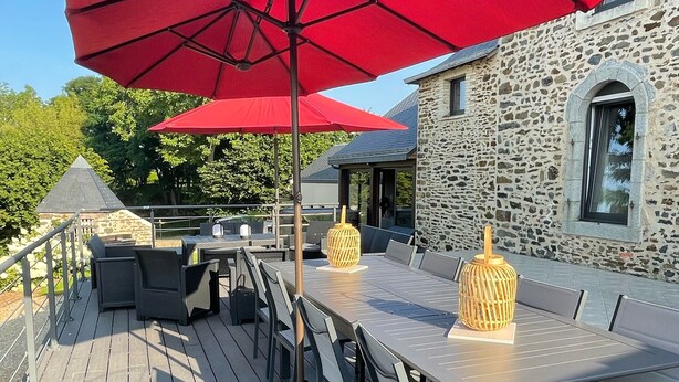 Une terrasse en bois avec une longue table et des chaises, des parasols rouges et un bâtiment en pierre en arrière-plan.
