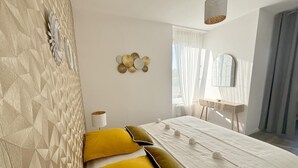 Une chambre moderne avec une tête de lit à motifs géométriques, un lit agrémenté d’oreillers jaunes et verts, une table de chevet avec une lampe, et un grand miroir accroché au mur.