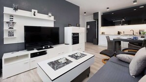 Una sala de estar moderna con televisor de pantalla plana, muebles blancos, una zona de comedor con una cocina al fondo y un sofá gris.
