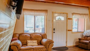A cozy room with a brown leather sofa, a white door, and a window with checkered curtains. - Aspen Ridge Cabin WIFI, 2bed+1Bath, 20mins to Yellowstone (Island Park)