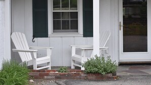 A white porch with two white Adirondack chairs, a window with green shutters, and a door with a glass panel.
