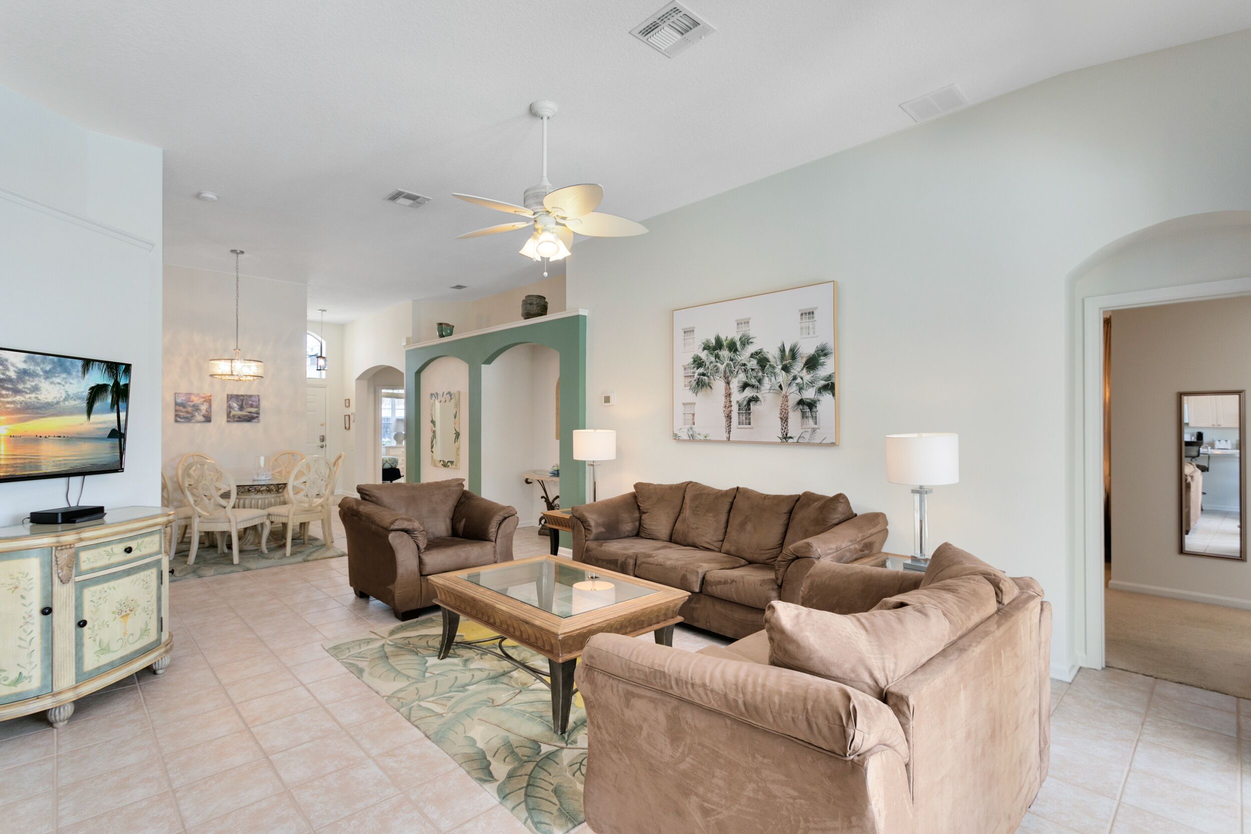 A spacious living room with a large brown sofa, a glass coffee table, and a television on a cabinet. There is a ceiling fan, a dining area with chairs, and a painting of palm trees on the wall.