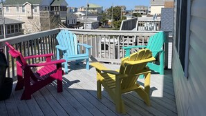 Una terraza de madera con sillas Adirondack de colores, barandilla y vistas a casas al fondo.
