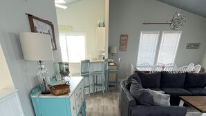 Amplia sala de estar con un sofá azul oscuro, un comedor con mesa azul claro y una zona de cocina al fondo.