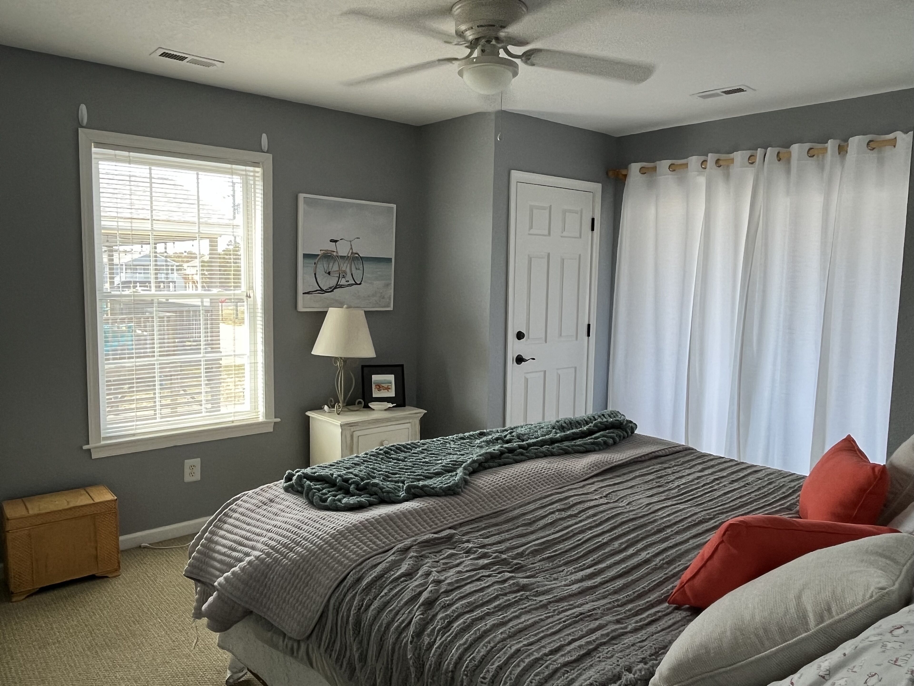 A bedroom with a bed, a nightstand, a lamp, a window with blinds, a door, and a ceiling fan.