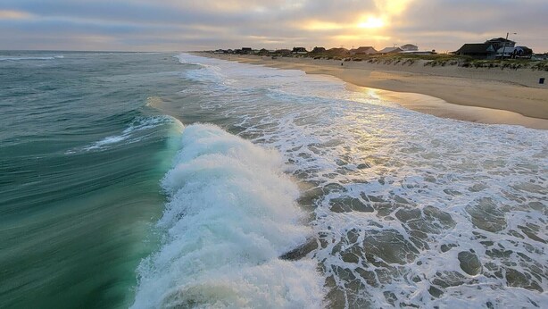 Una playa con olas rompiendo en la orilla, un atardecer al fondo y edificios a lo largo de la costa.