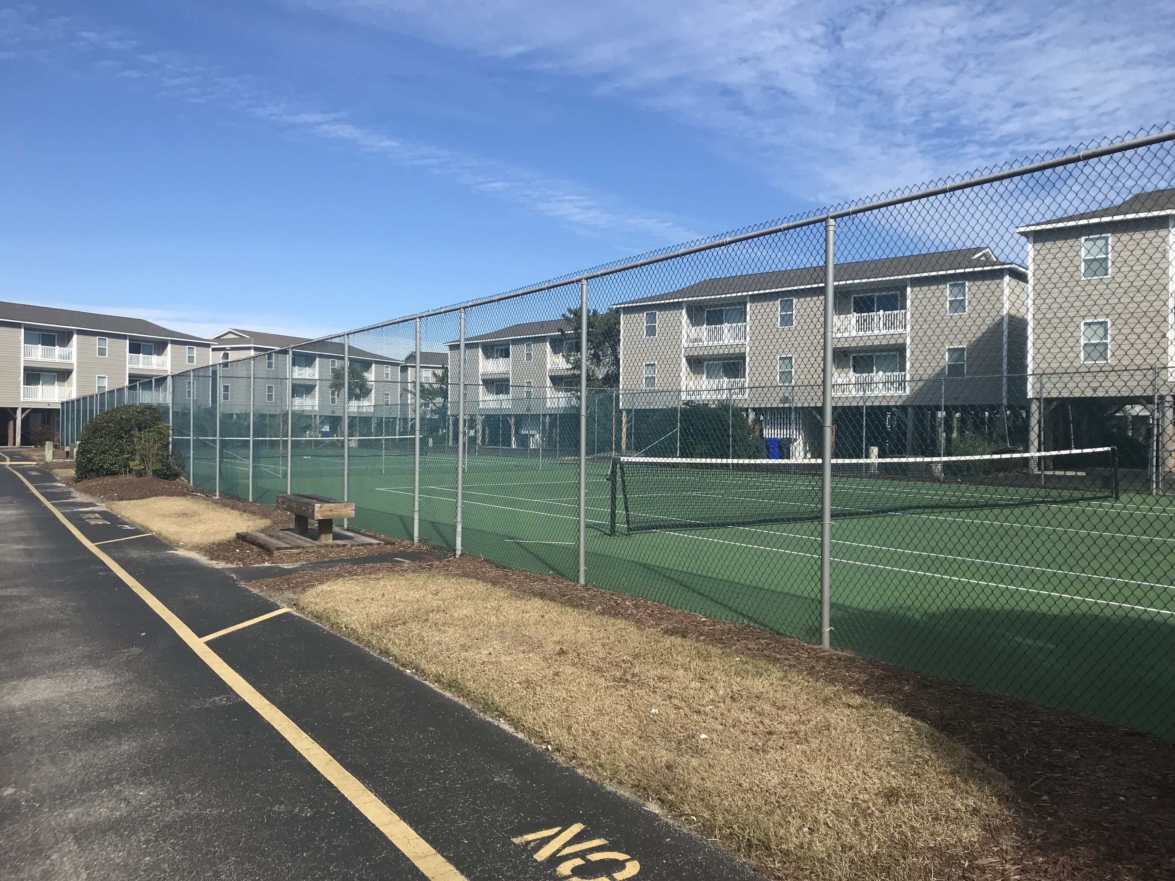 A tennis court surrounded by a chain-link fence, with a parking lot and residential buildings in the background.