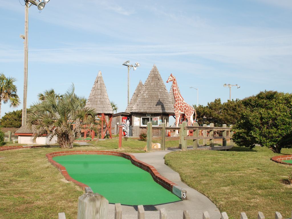 Un campo de mini golf con un área de juego verde, estructuras de madera que parecen chozas y una gran estatua de jirafa.