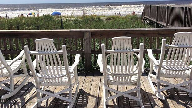 Une terrasse en bois avec des fauteuils Adirondack blancs donnant sur une plage où se trouvent des personnes et des parasols.